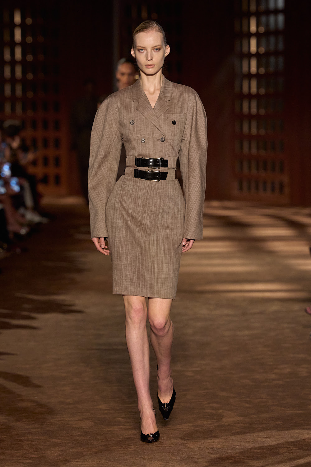 Modelo con un vestido sastre de manga larga y hombreras pronunciadas en tono marrón claro, ajustado en la cintura con un doble cinturón de piel negra con hebillas doradas, cortesía de Ferragamo.