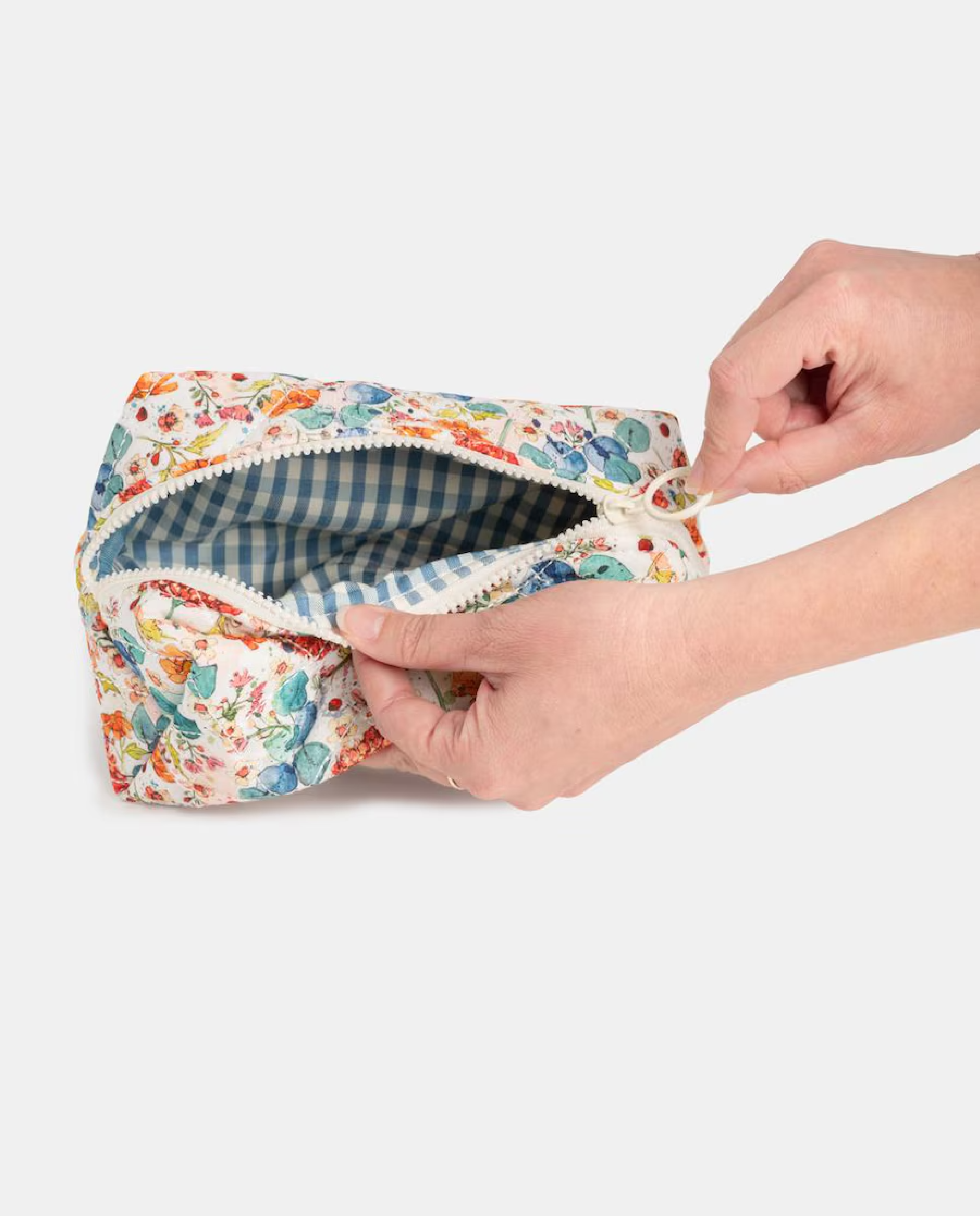Primer plano de manos abriendo un neceser de tela con estampado floral multicolor y un forro interior de cuadros vichy azul y blanco.