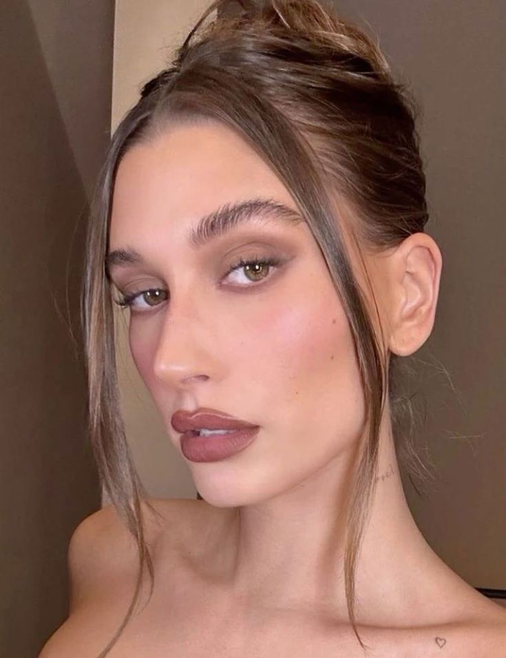 Primer plano del rostro de una mujer con el cabello recogido, luciendo un maquillaje en tonos tierra cálidos con sombras ahumadas sutiles en los párpados y labios delineados en tono marrón.