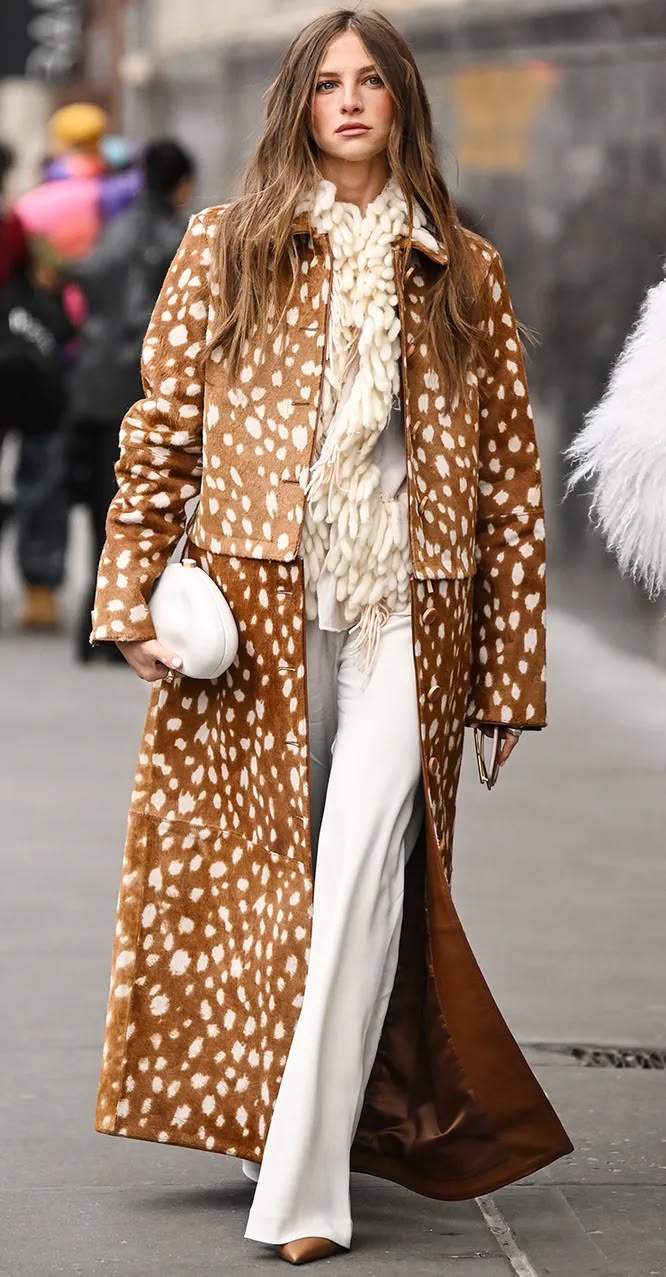 Mujer desfilando por la calle con un abrigo largo con estampado de bambi, pantalones blancos fluidos, una bufanda de lana texturizada y un bolso de mano blanco estructurado.