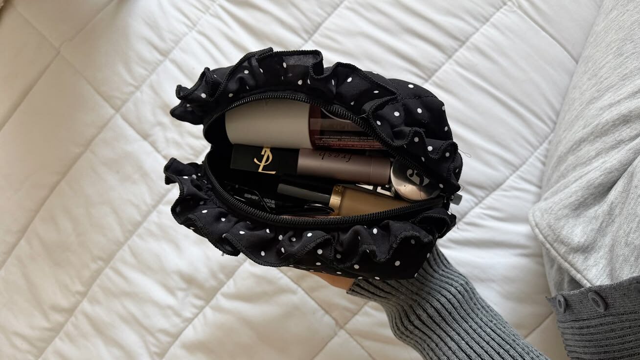 Vista superior de un neceser negro de tela con estampado de lunares blancos y bordes con volantes fruncidos, abierto y mostrando en su interior varios productos de maquillaje y cosmética como un labial de Yves Saint Laurent y un bálsamo labial.