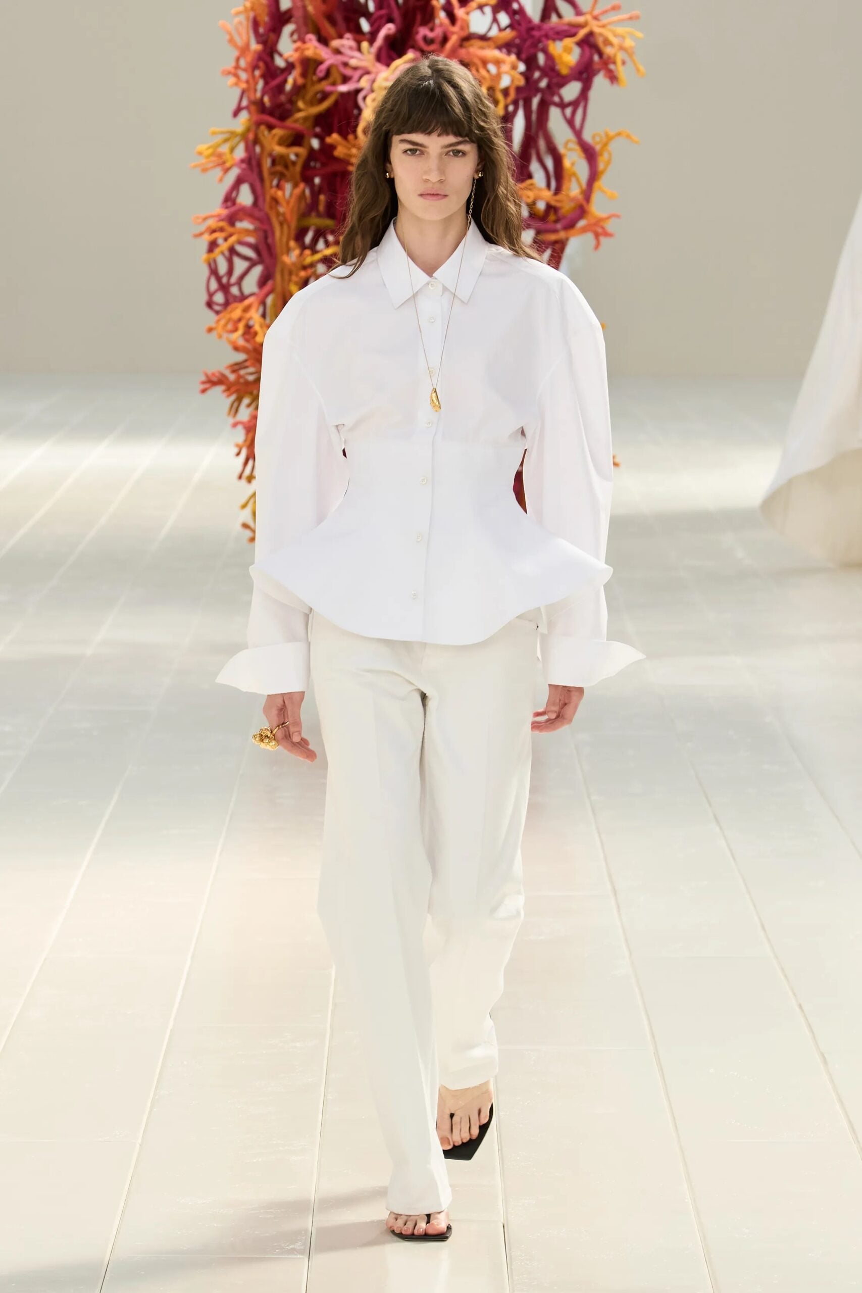 Modelo desfilando con una camisa blanca de manga larga con hombreras marcadas, peplum estructurado en la cintura y pantalones blancos rectos, complementada con un collar largo dorado y sandalias oscuras