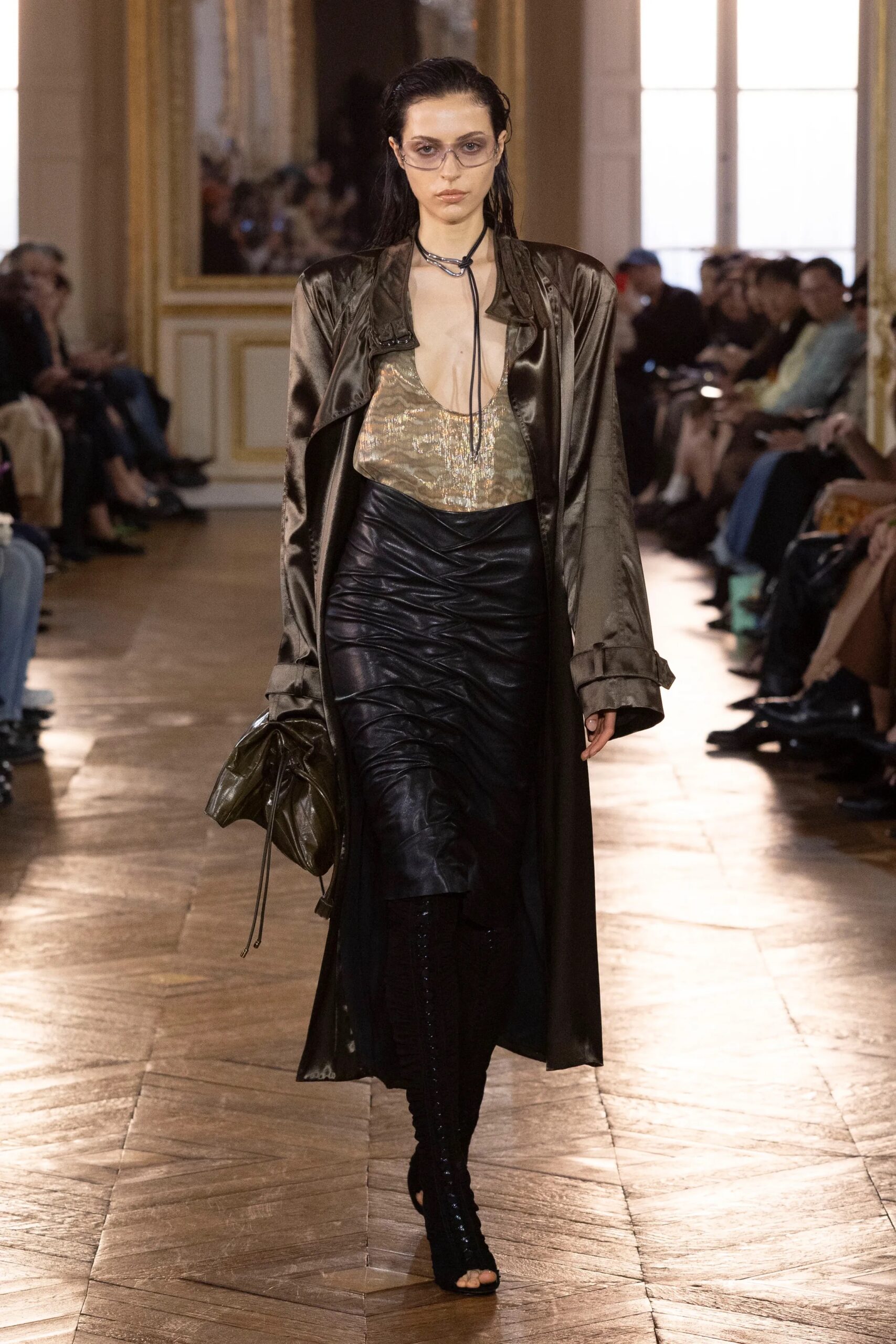 Modelo desfilando con una gabardina larga satinada en tono bronce, un top metalizado brillante, falda lápiz negra efecto piel con drapeados frontales y botas altas oscuras.