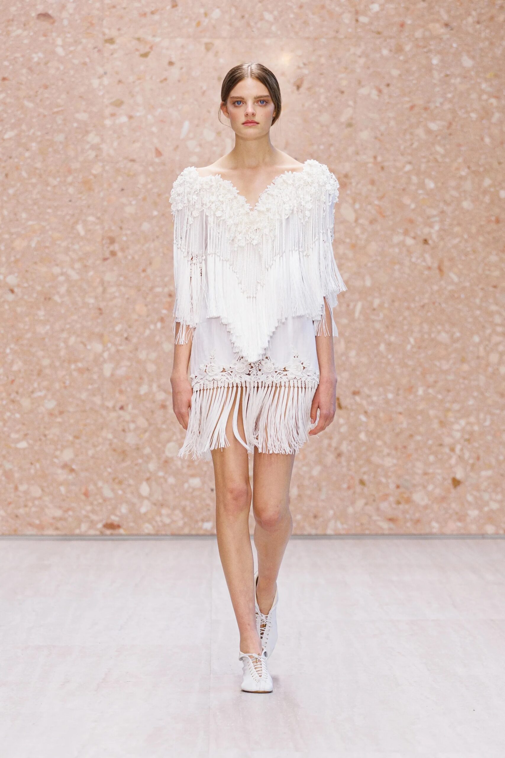 Modelo desfilando con un minivestido blanco de escote en V pronunciado, cubierto con una capa de flecos largos y bordados florales texturizados, complementado con zapatos bajos acordonados de piel blanca.