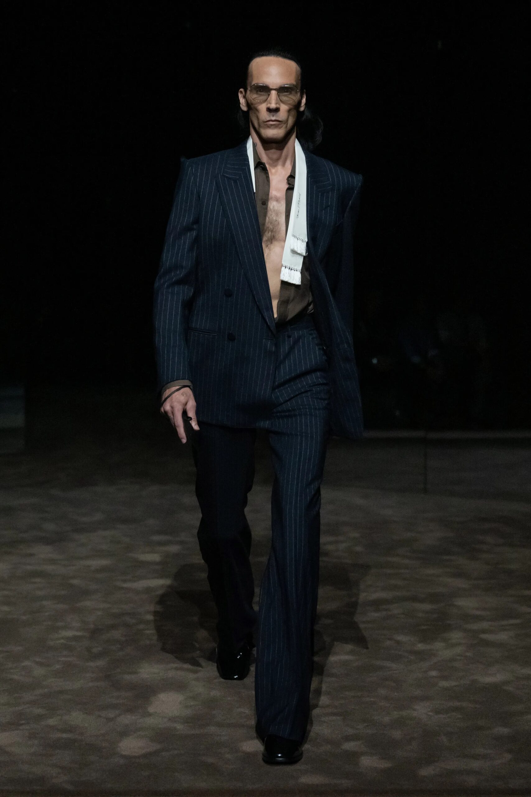 Modelo masculino desfilando con un traje cruzado de raya diplomática en tono oscuro, camisa marrón abierta al pecho y una bufanda blanca ligera colgada al cuello.