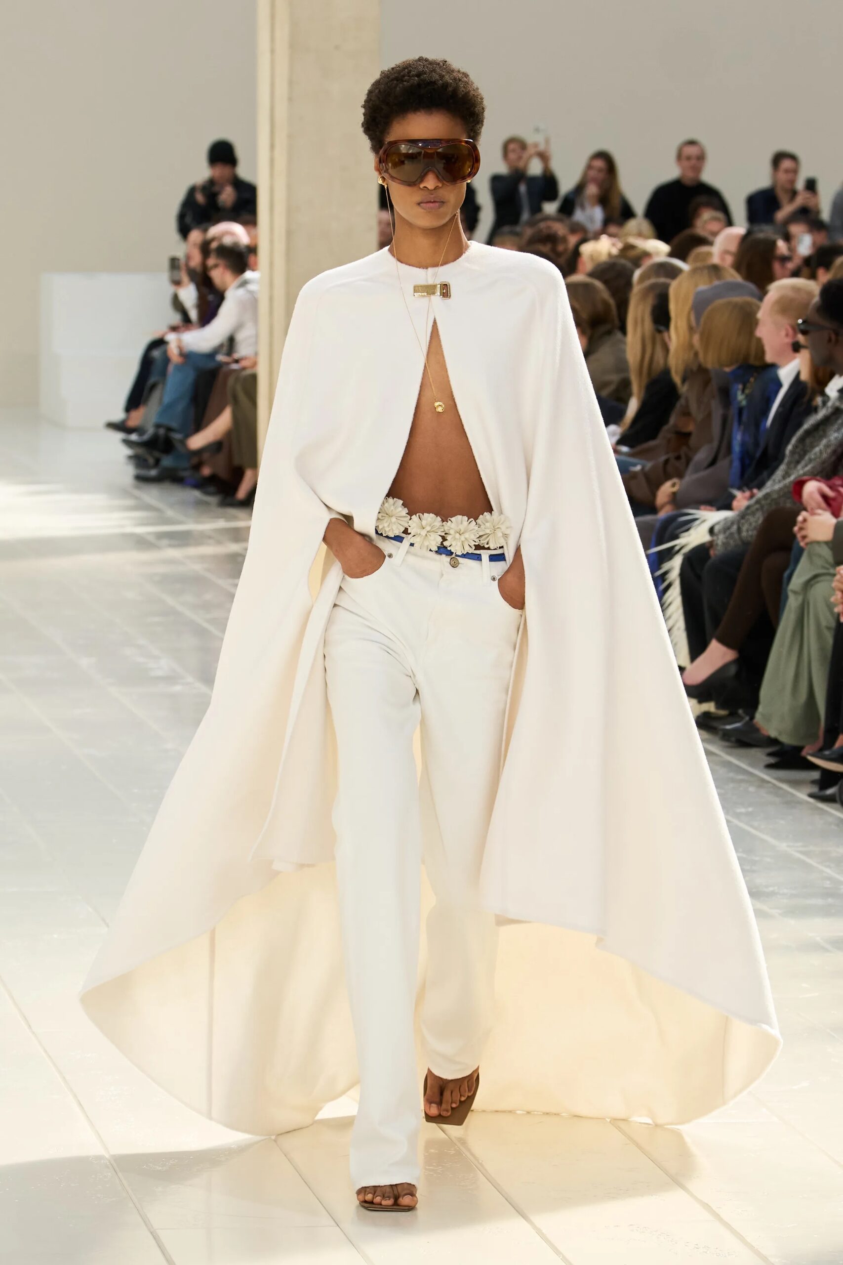 Modelo portando una capa blanca larga y fluida con una abertura frontal, pantalones blancos con un cinturón decorado con motivos florales, gafas de sol envolventes y un collar delicado.
