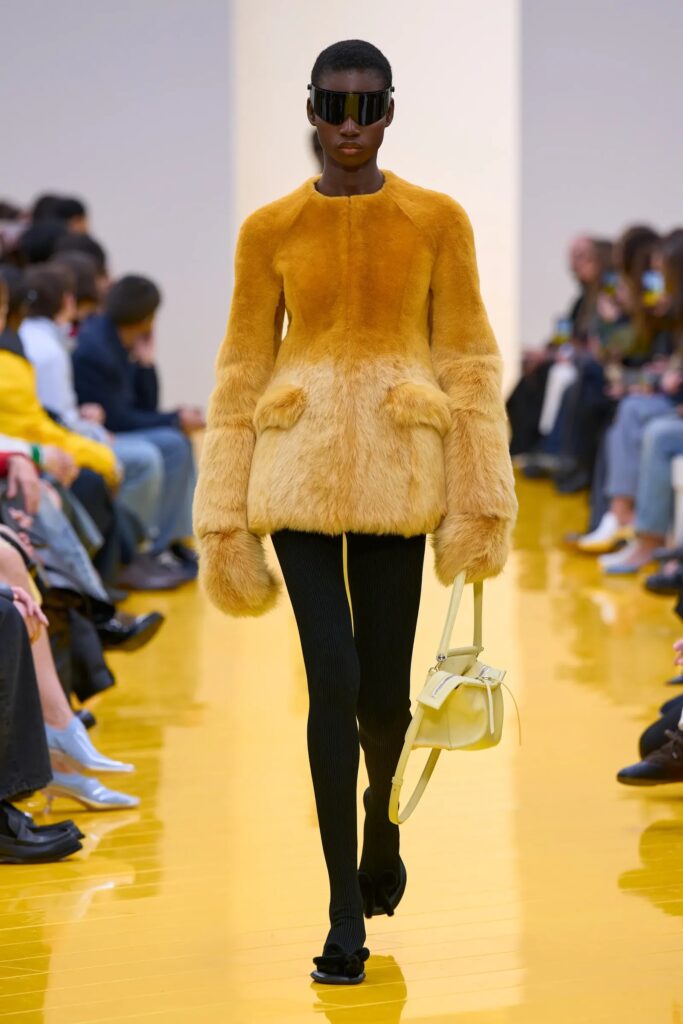 Modelo desfilando con un abrigo de peluche amarillo mostaza, mallas negras y sosteniendo una bolsa de hombro pequeña de color amarillo pálido