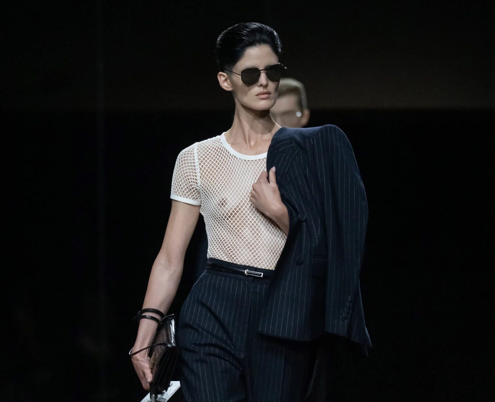 Modelo portando un pantalón sastre oscuro de raya diplomática con cinturón, una camiseta ajustada de red blanca semitransparente, gafas de sol y llevando una chaqueta y un maletín en la mano.