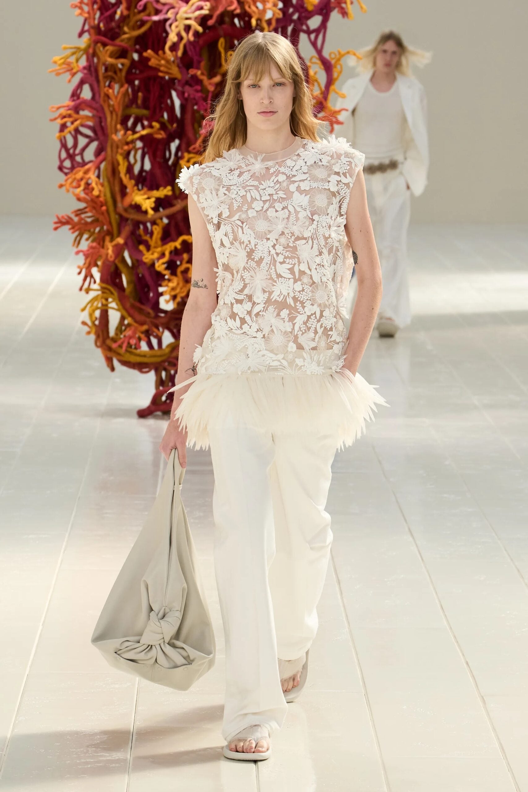 Modelo luciendo una blusa blanca sin mangas con intrincados bordados florales transparentes y un borde inferior de plumas, combinada con pantalones blancos rectos, sandalias y un bolso de mano suave.