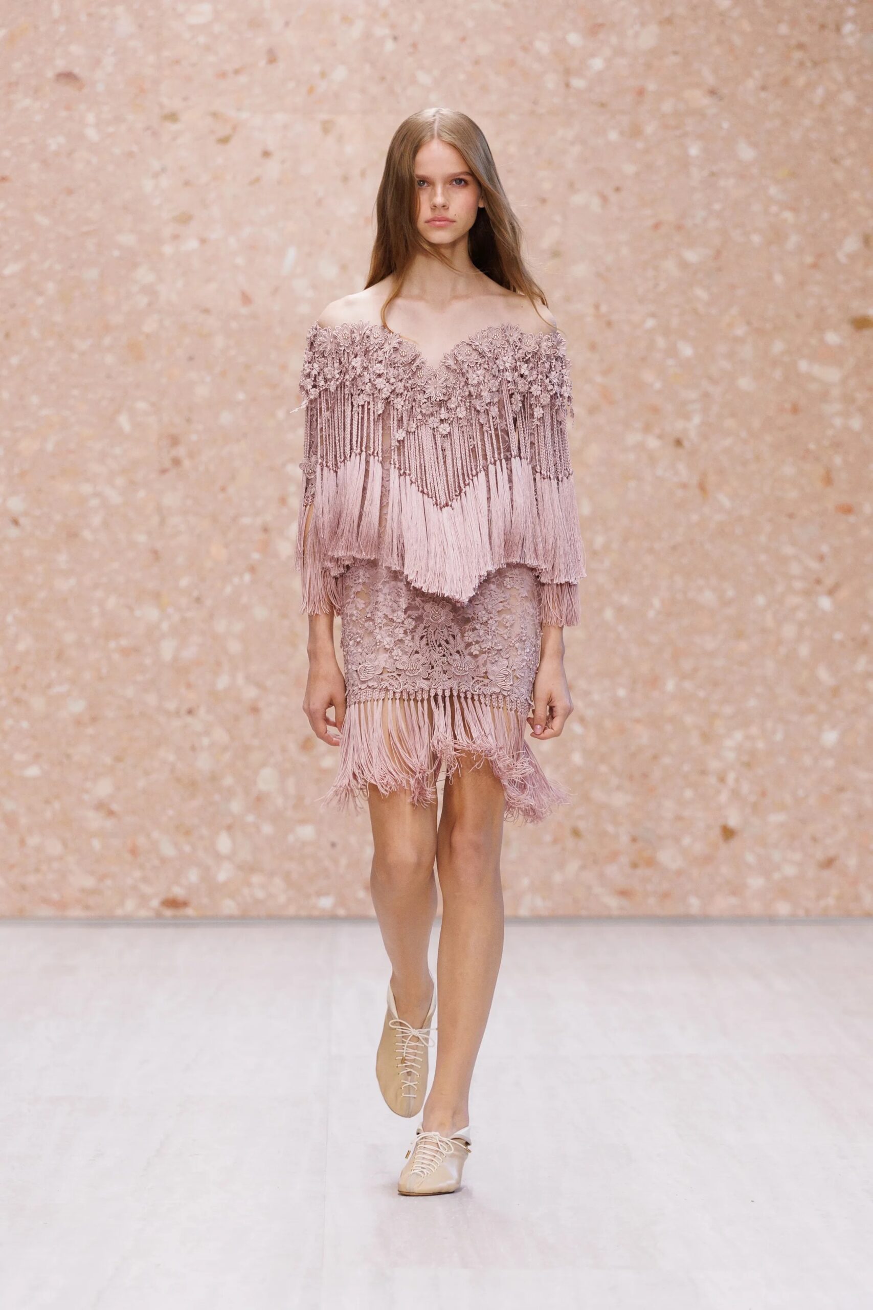 Modelo luciendo un conjunto en tono rosa polvoriento, compuesto por una blusa de hombros caídos con flecos y aplicaciones florales, un vestido corto ajustado de encaje y calzado plano en tonos tierra.