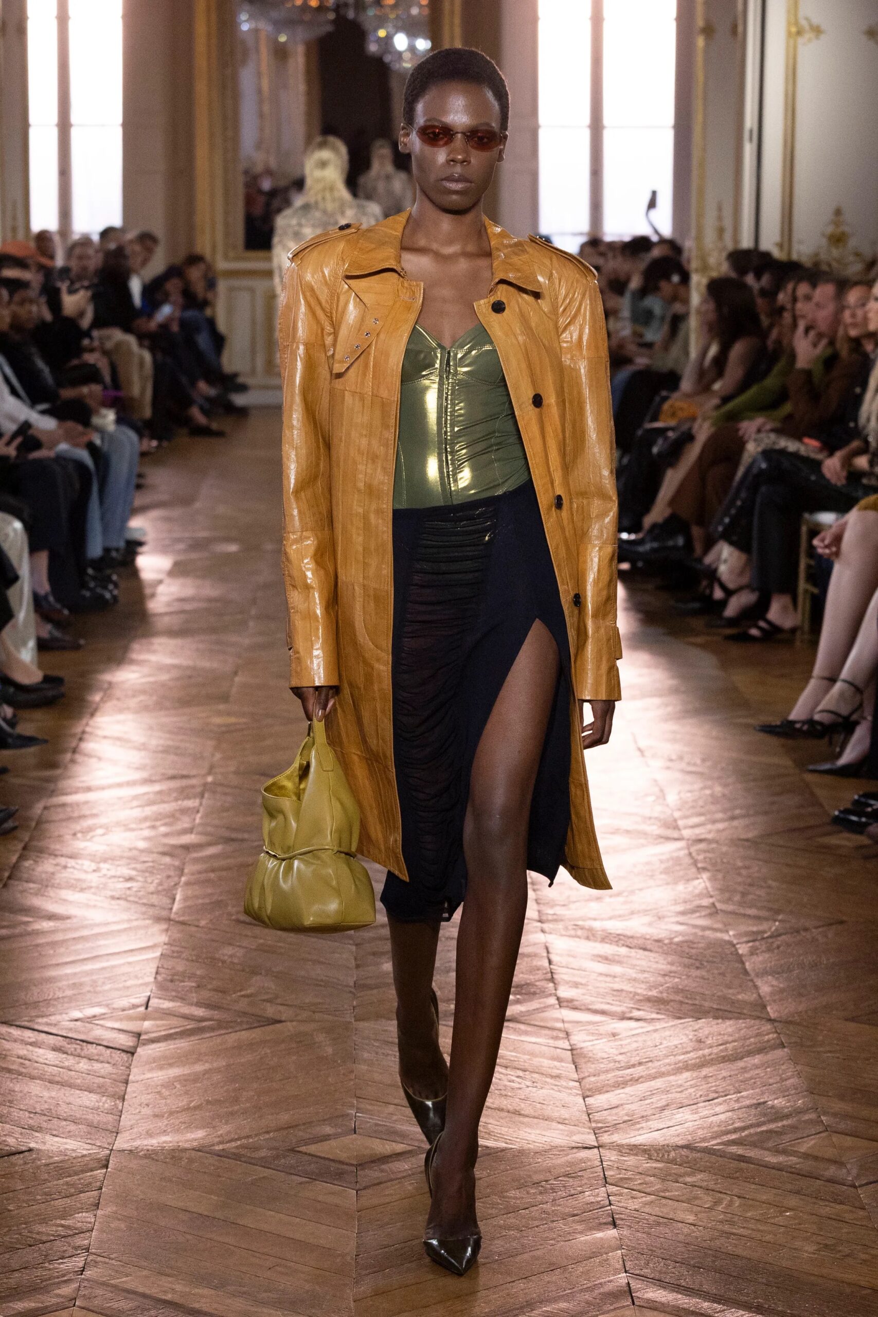 Modelo caminando por la pasarela con un abrigo largo de cuero en tono ocre brillante, un corpiño metálico dorado y una falda midi negra con abertura pronunciada.