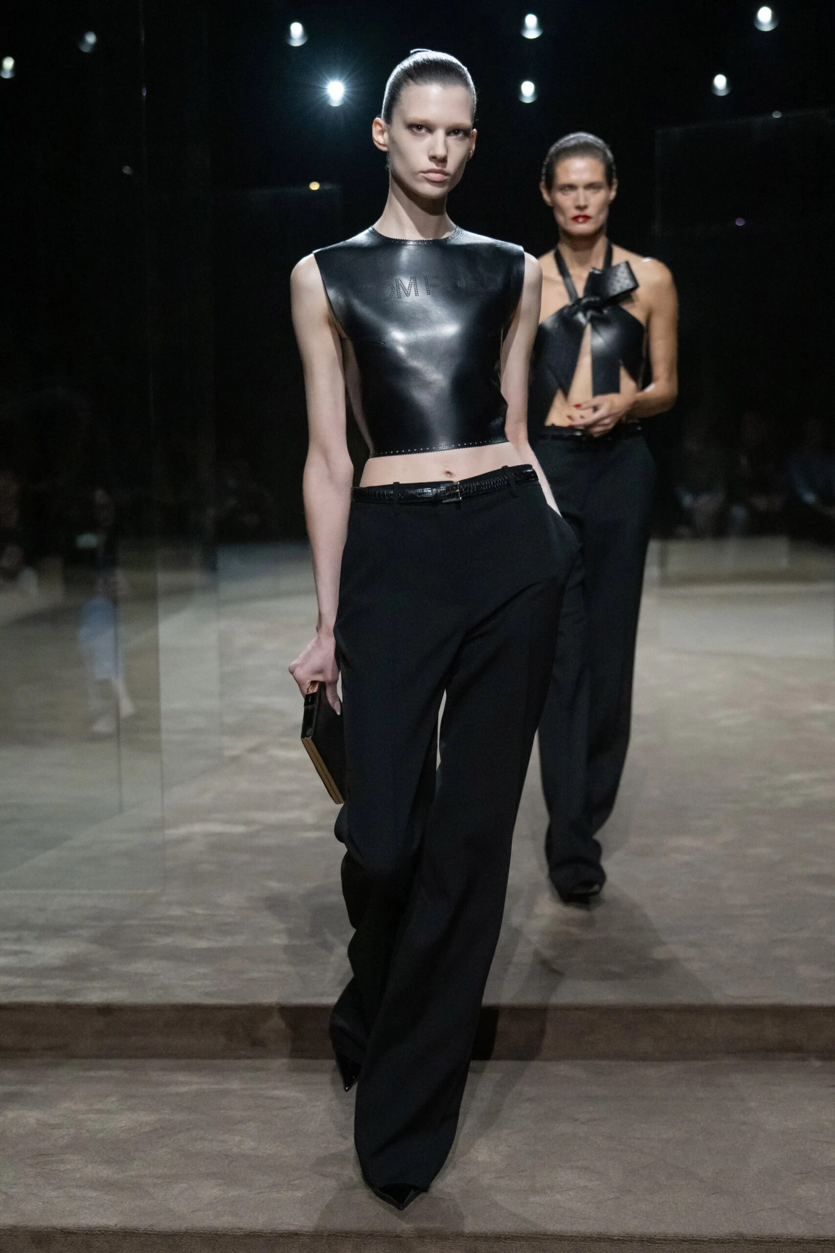Modelo luciendo un top corto tipo corsé de piel sintética negra sin mangas con el cuello alto, combinado con pantalones negros fluidos de talle alto y un bolso de mano rígido.