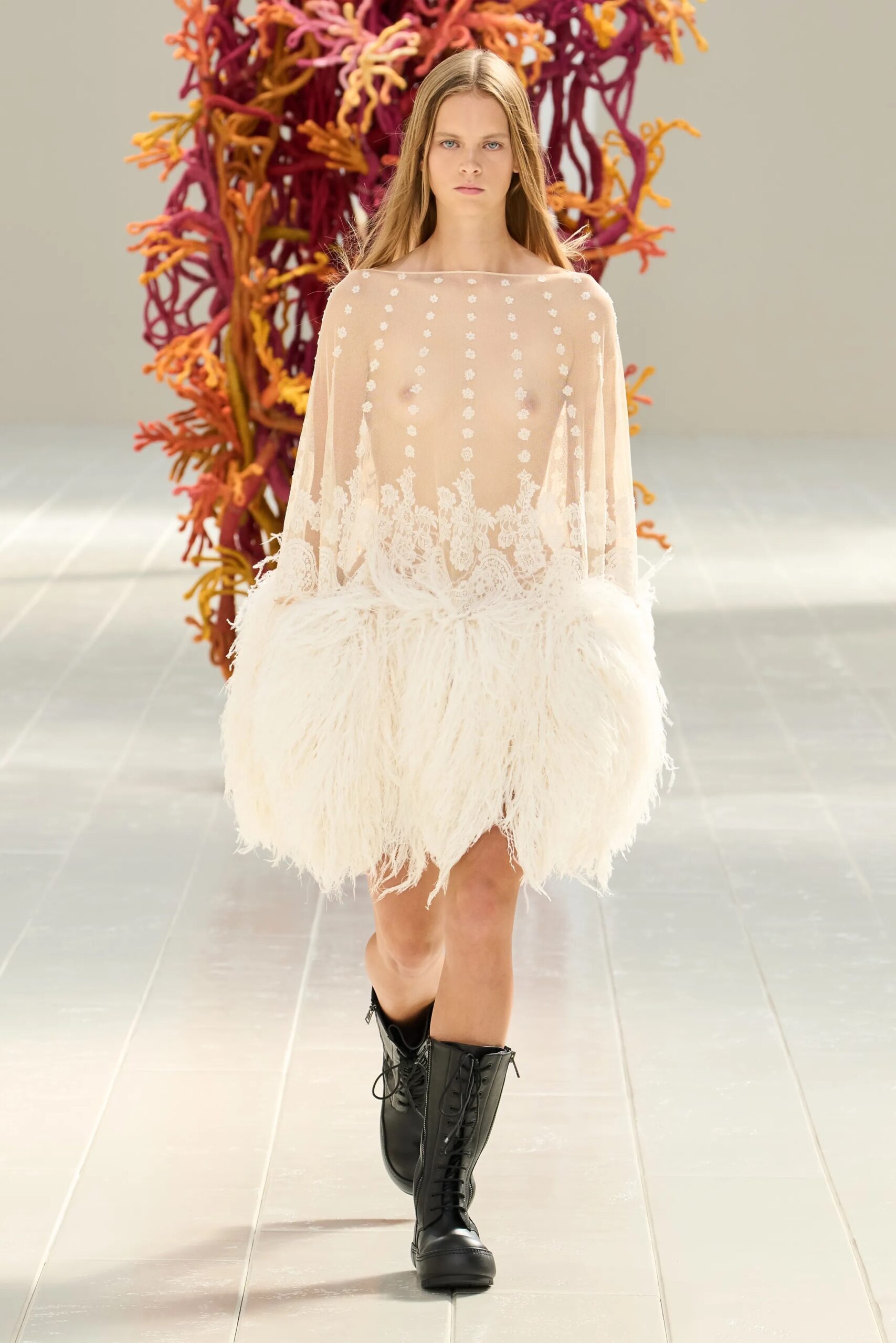 Modelo portando un vestido corto translúcido en tono marfil con detalles bordados sutiles y un bajo voluminoso de plumas, combinado con botas altas negras con cordones.