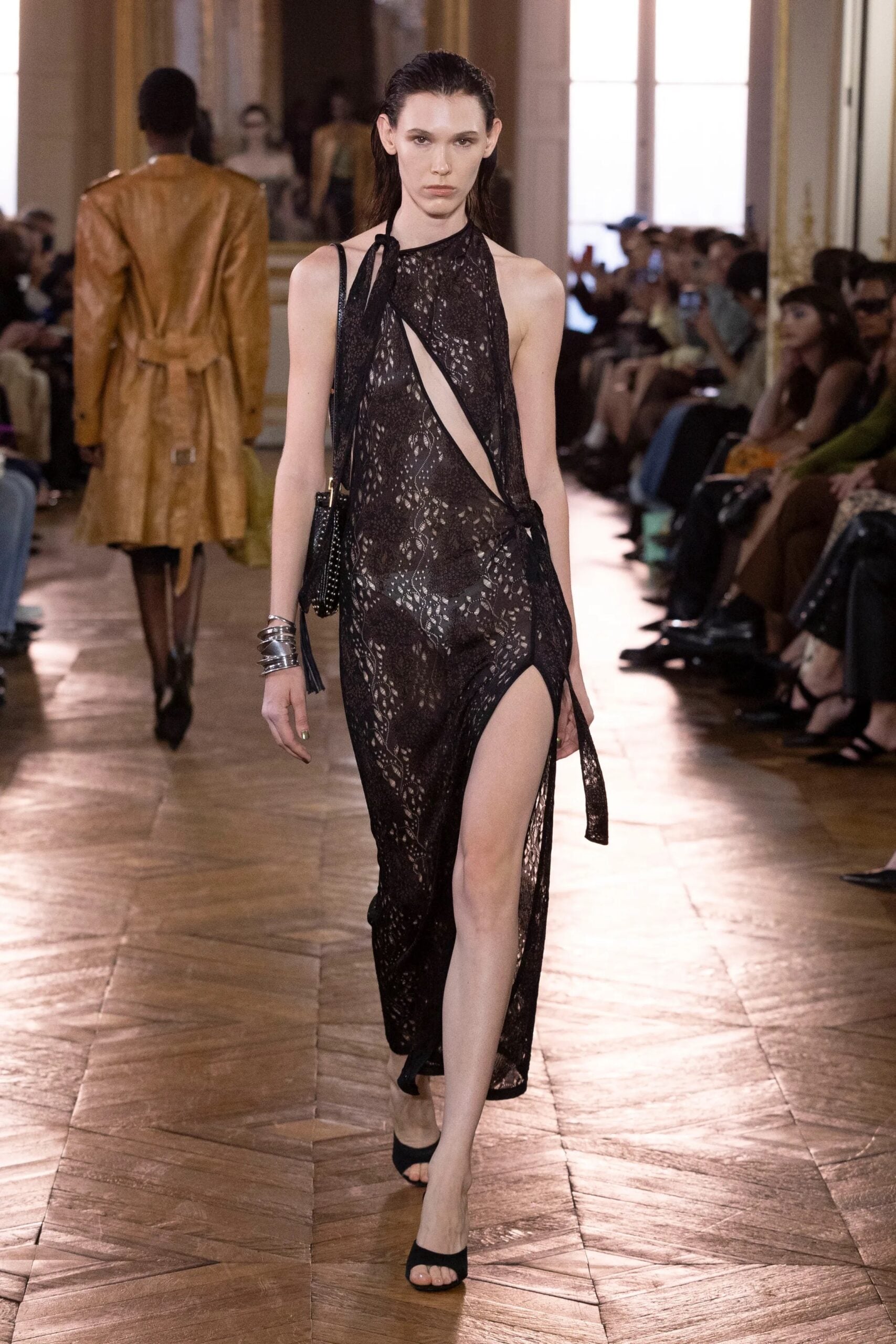 Modelo desfilando con un vestido largo de encaje negro con aberturas diagonales asimétricas y una profunda abertura en la pierna, complementado con pulseras metálicas plateadas.