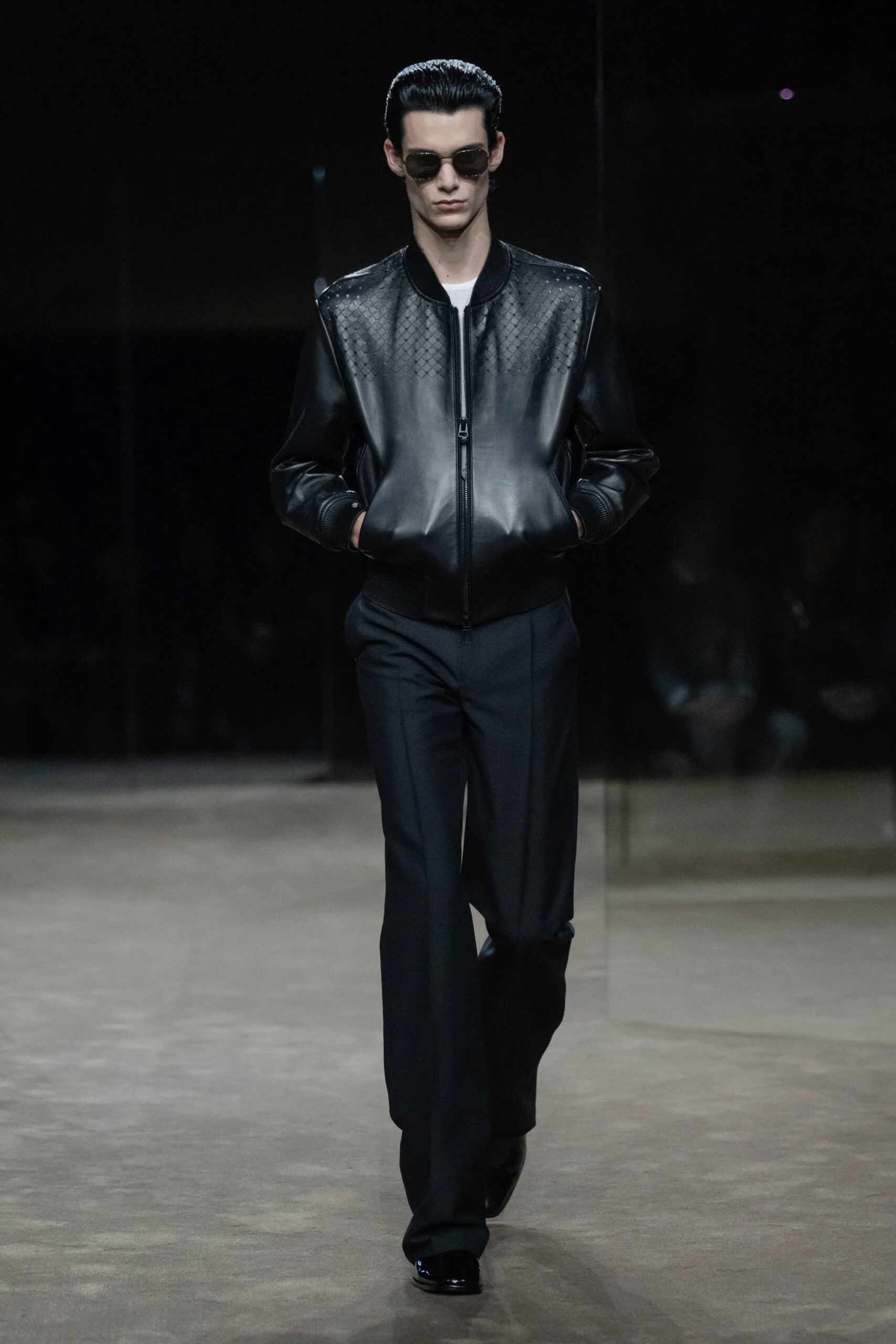 Modelo desfilando con una chaqueta bomber de cuero negro con paneles texturizados, pantalones oscuros de corte recto y gafas de sol oscuras.