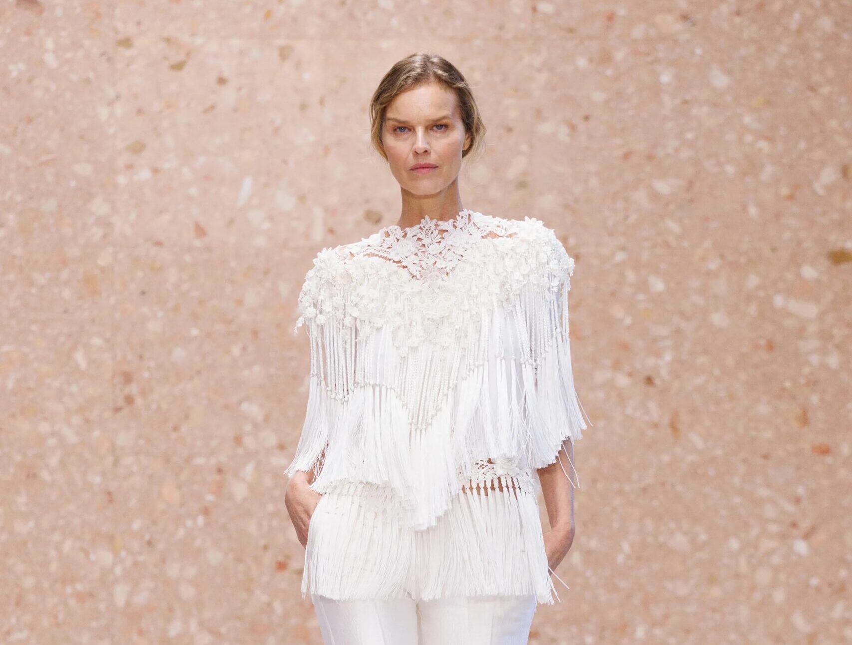 Modelo vistiendo una blusa blanca estructurada con bordados de encaje en el cuello y cascadas de flecos, combinada con bermudas sastre blancas y sandalias minimalistas de tacón fino.