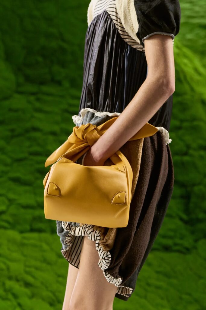 Detalle de una modelo sosteniendo una bolsa de mano amarilla suave con un detalle de nudo, sobre un atuendo de capas oscuras con texturas