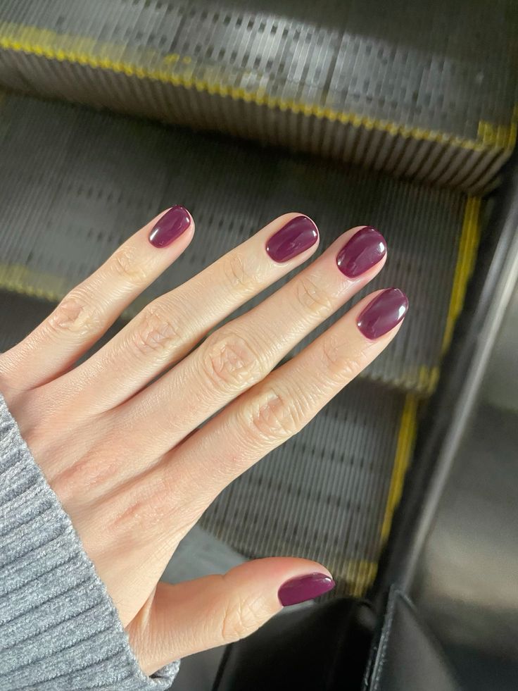 Mano estirada mostrando una manicura corta en un tono ciruela ligeramente más rojizo y vibrante, con el fondo metálico de unas escaleras mecánicas.