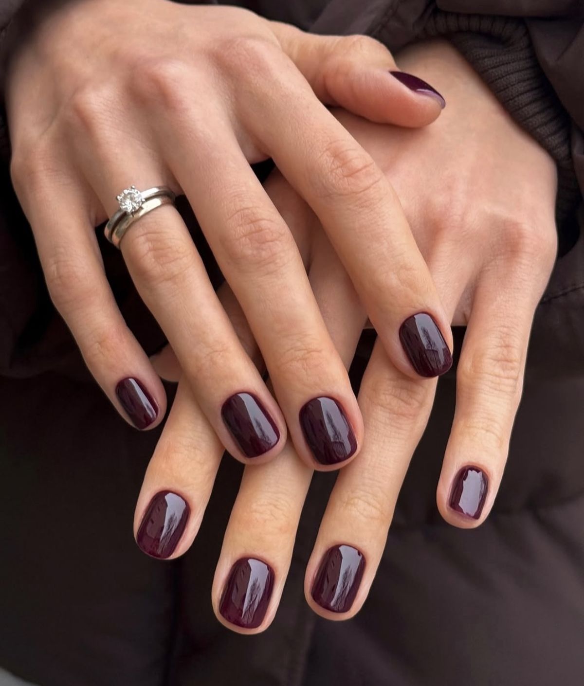Mano con manicura en tono ciruela oscuro posada elegantemente sobre una superficie de mármol junto a ciruelas frescas y maduras.