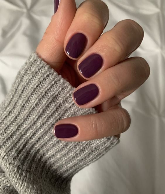 Mano con uñas cortas de forma almendrada suave en un elegante color ciruela berenjena, luciendo un suéter de punto gris claro.