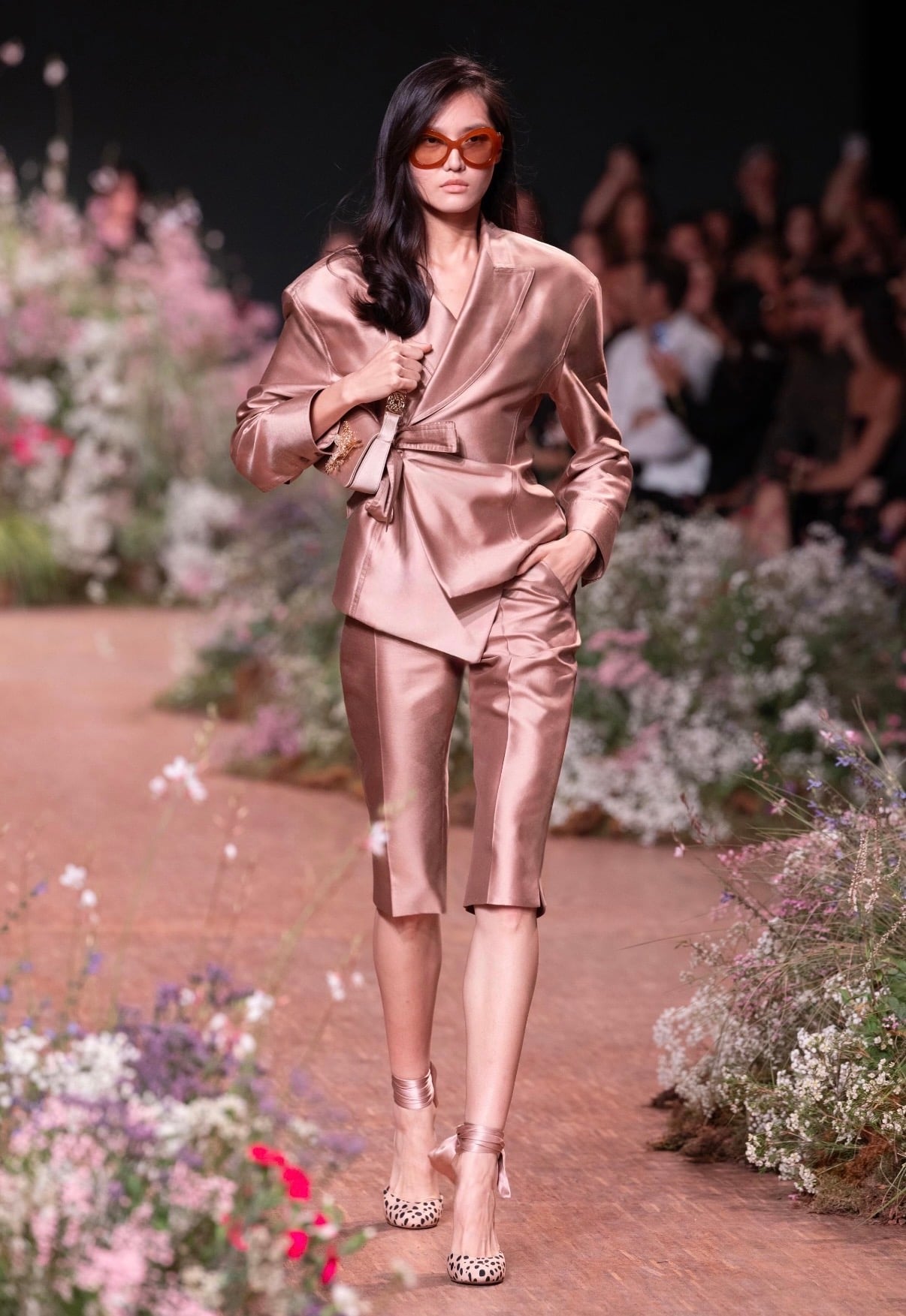 mujer en desfile con cabello negro de lado y lentes. Con saco y capri pants rosas de seda