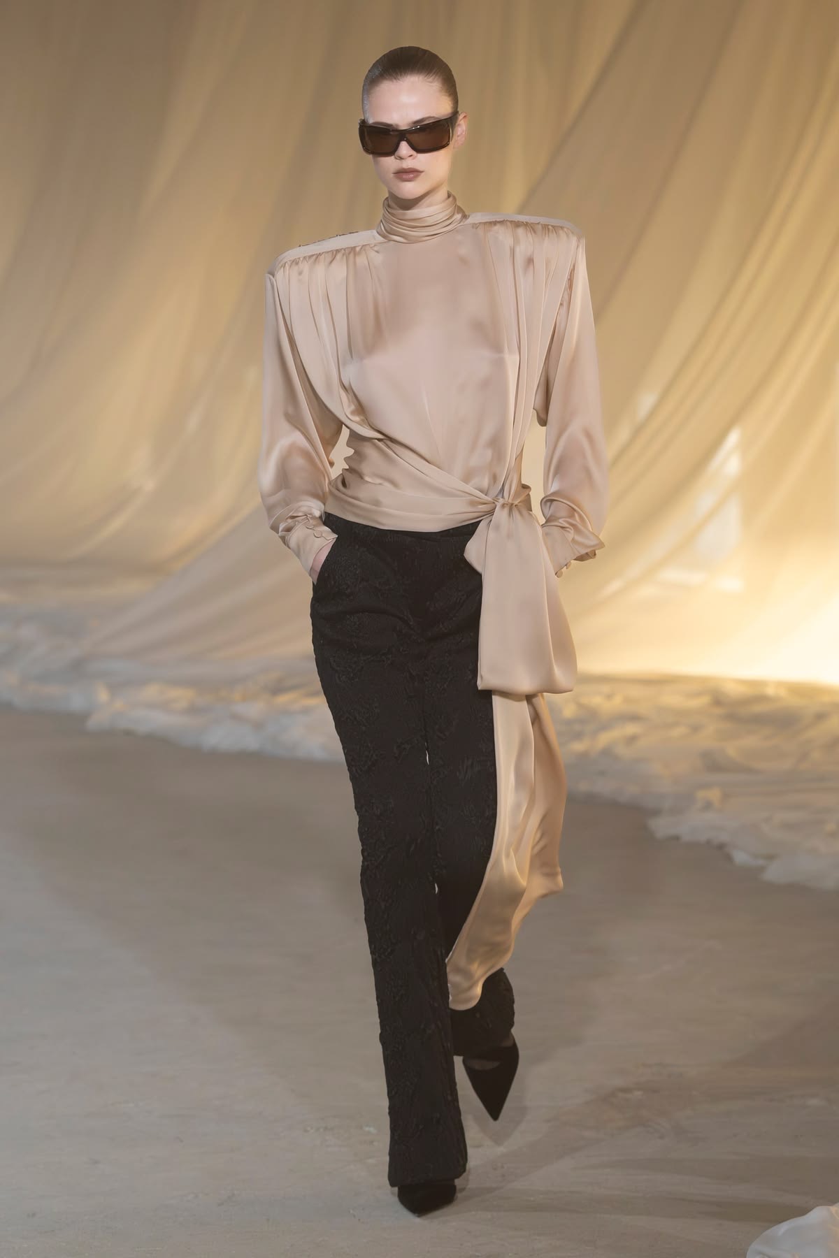 modelo de lentes obscuros con blusa beige drapeada con lazo de lado y pantalones negros