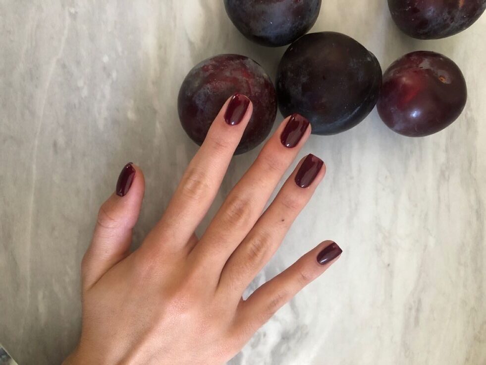 Manos cruzadas mostrando una manicura corta y redondeada en un tono ciruela oscuro, brillante y pulido, con un anillo plateado con diamante en el dedo anular.