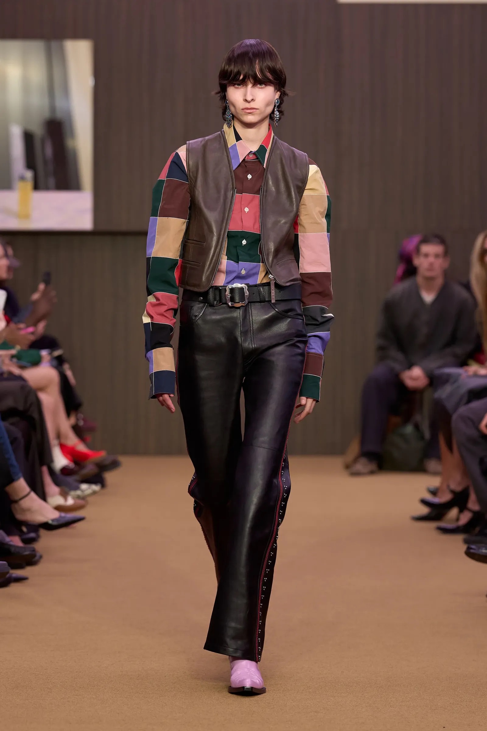 Modelo desfilando con una camisa de patchwork multicolor, chaleco de cuero, pantalones oscuros y unos zapatos de tacón en tono lila suave.