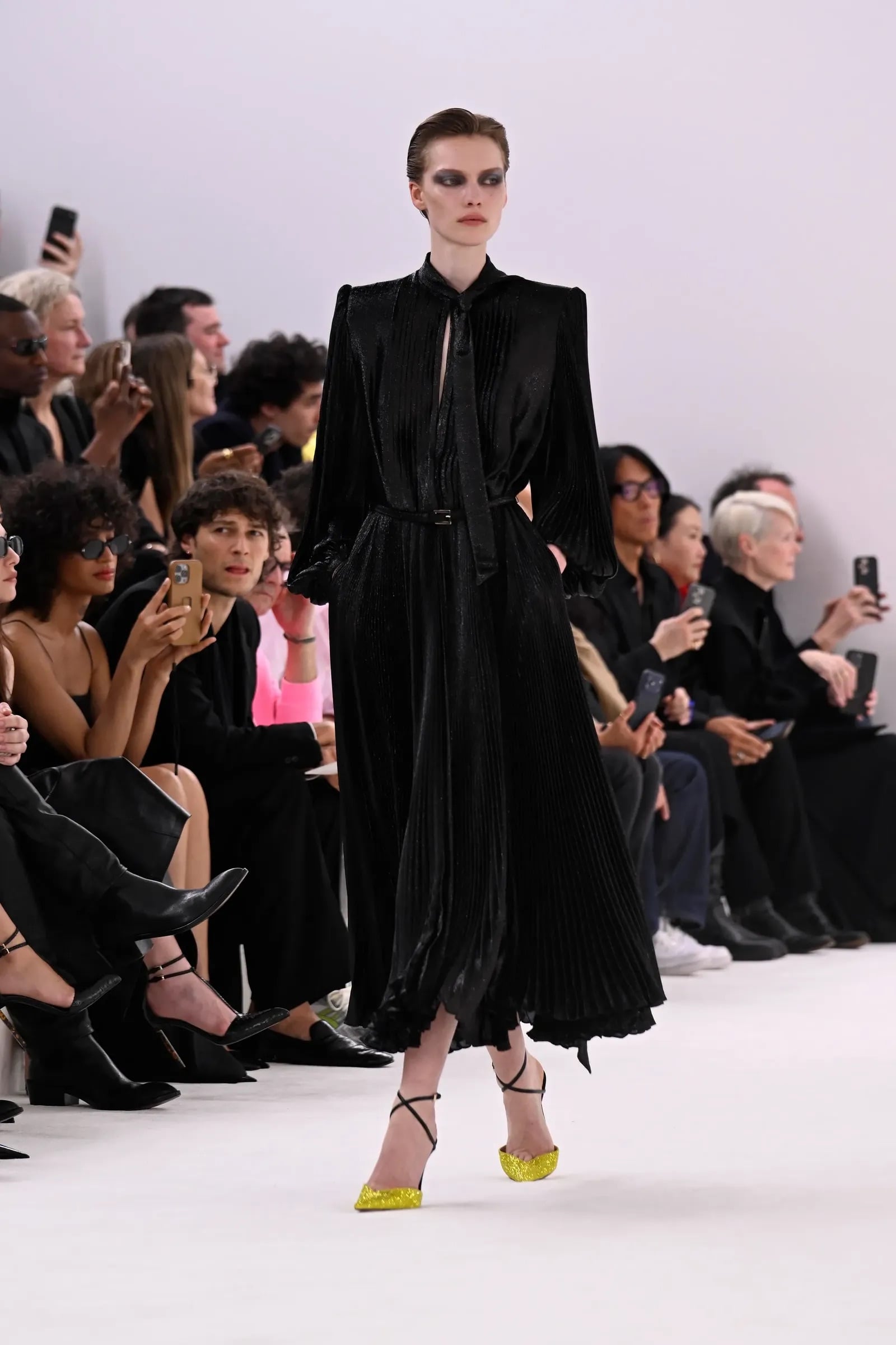 Modelo caminando con un vestido negro plisado de manga larga y unos zapatos de tacón destalonados en un llamativo color amarillo brillante con acabado brillante.
