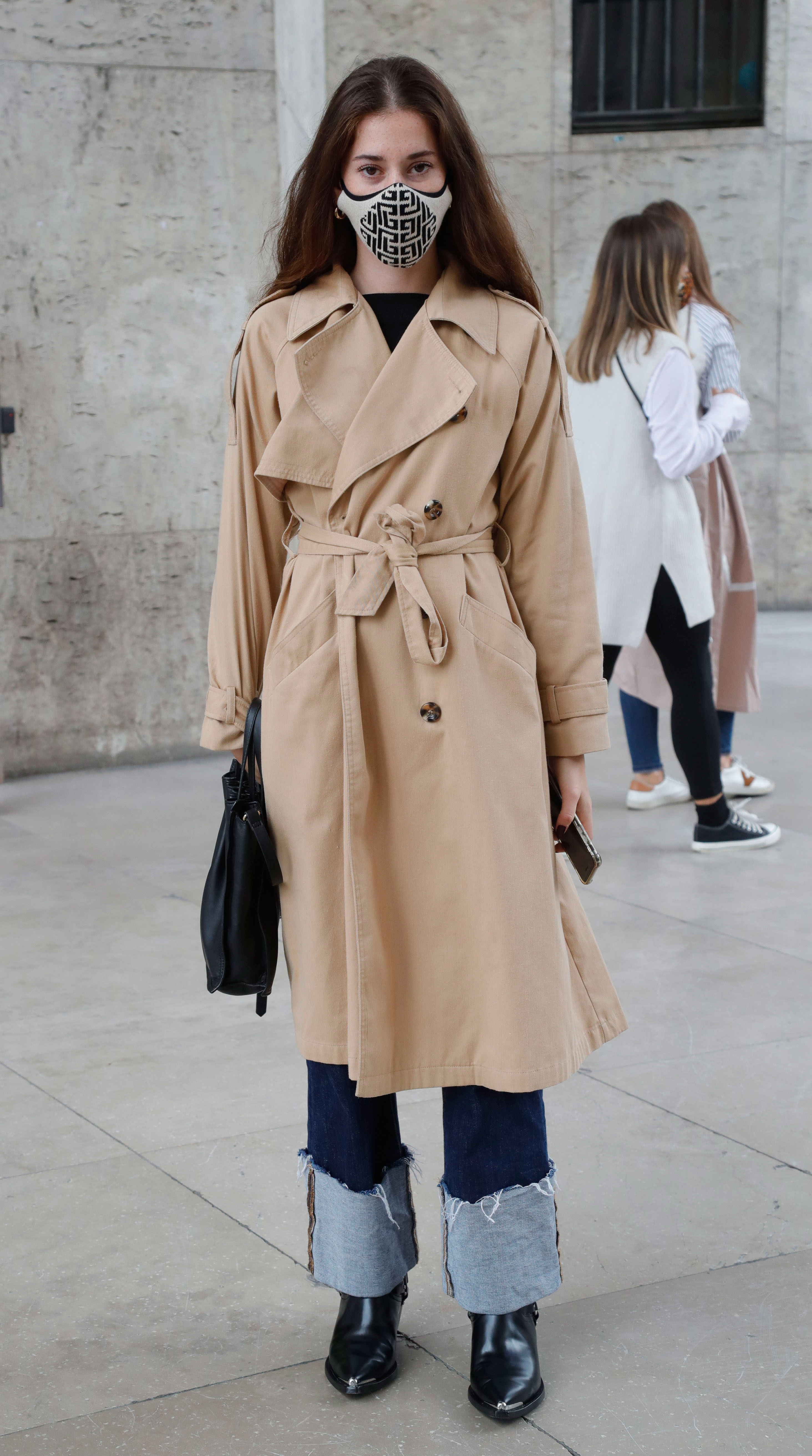 Paris Fashion Week Street Style: Woman wears a trench coat, mask, jeans, and black boots.