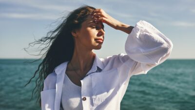 Romanticize Your Life Psychology. Photo via Getty Images. Woman standing by the sea with hand on forehead. Hair blowing in the wind. Clear sky