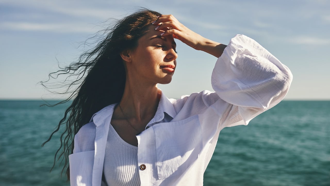 Romanticize Your Life Psychology. Photo via Getty Images. Woman standing by the sea with hand on forehead. Hair blowing in the wind. Clear sky