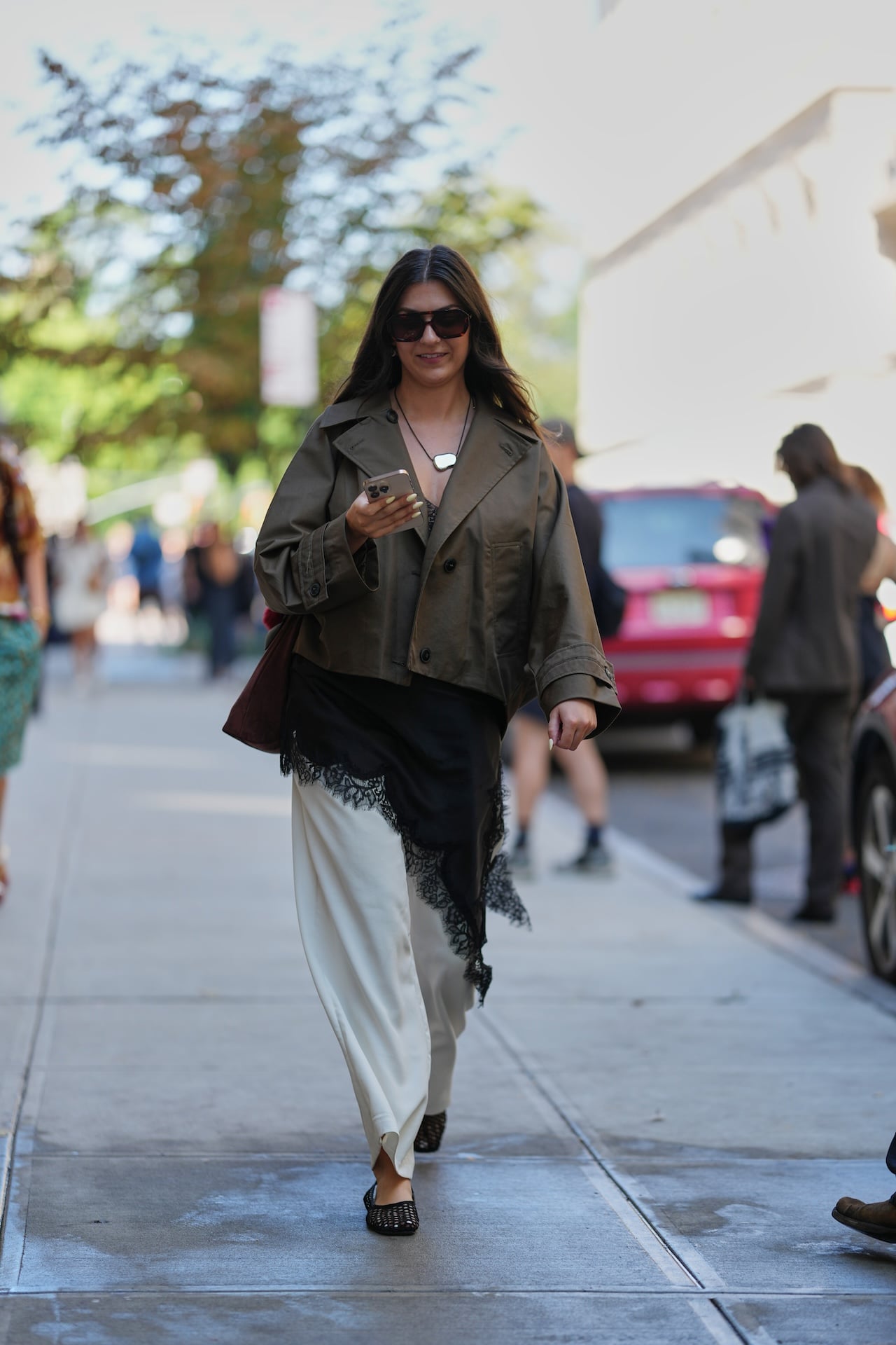 NEW YORK, NEW YORK - SEPTEMBER 14: A guest wears long dark brown hair worn loose, oversized dark brown rectangular sunglasses, small gold hoop earrings and a black pendant necklace with a white irregular-shaped pendant on a black cord, a soft burgundy suede shoulder bag, an olive-green cropped cotton trench coat with wide lapels and black buttons, a black silk slip with scalloped black lace trim worn asymmetrically over cream satin wide-leg trousers, black woven leather flat shoes, outside Ulla Johnson, during New York Fashion Week, on September 14, 2025 in New York, New York (Photo by Edward Berthelot/Getty Images)