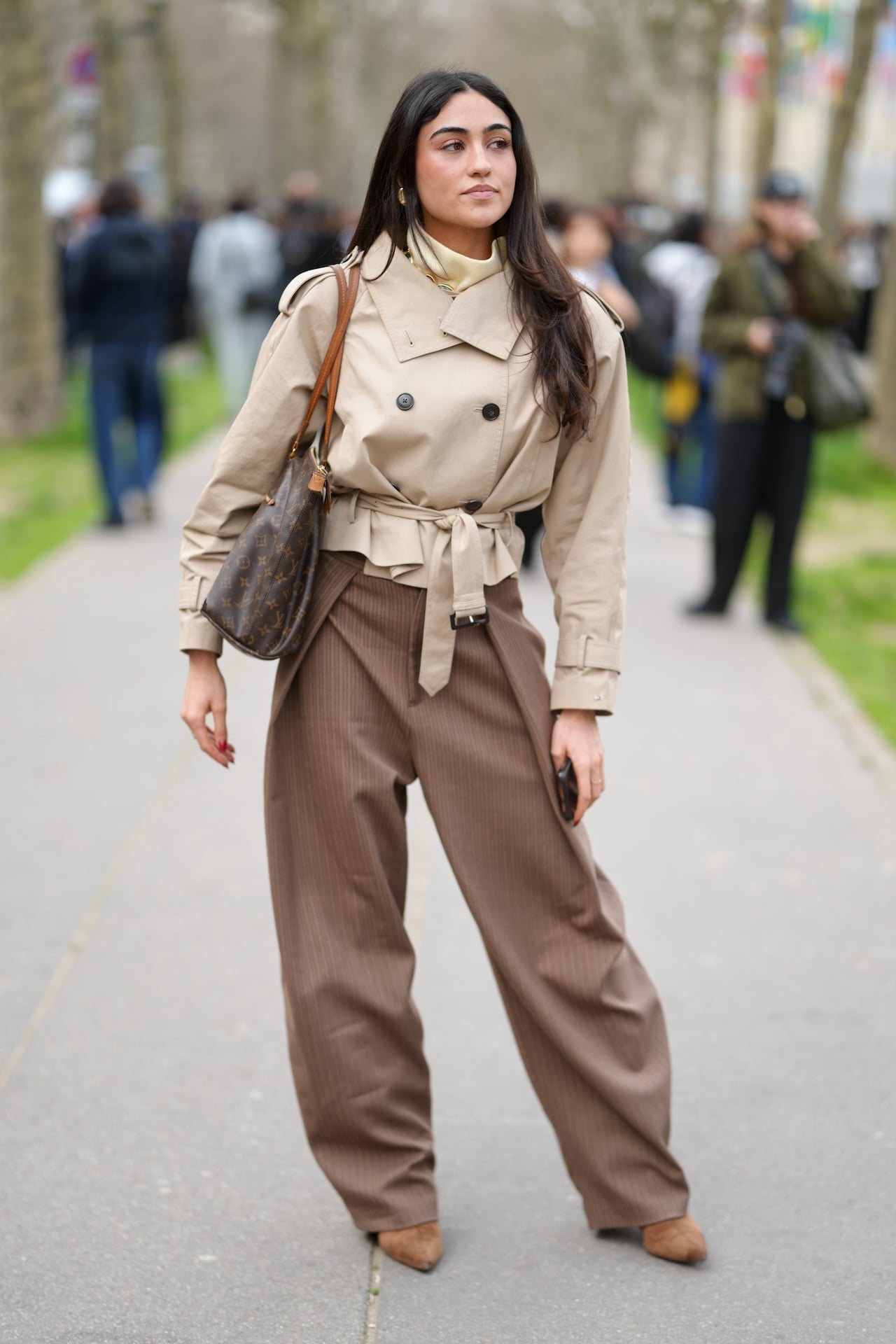 PARIS, FRANCE - MARCH 05: A person is seen wearing gold hoop earrings, a cream turtleneck, a beige cropped trench coat with a belted waist, brown pinstripe wide-leg trousers, tan pointed-toe boots, while carrying a Louis Vuitton brown shoulder bag during Day Four of Paris Fashion Week - Womenswear Fall/Winter 2026/2027 on March 05, 2026 in Paris, France. (Photo by 305pics/Getty Images)