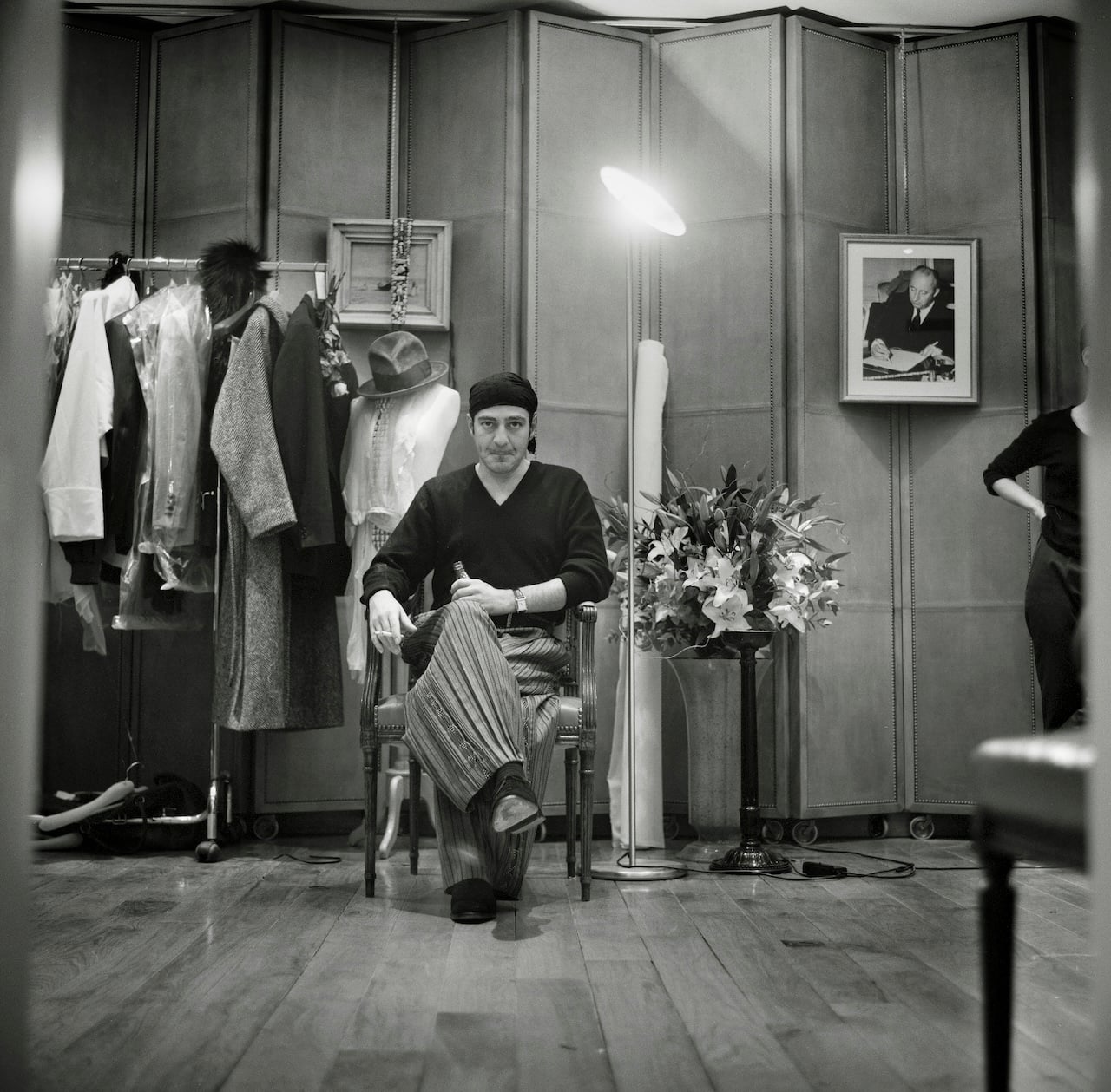 PARIS, FRANCE - 2005: British fashion designer John Galliano wearing baggy striped trousers, a black bandana head covering and a V-neck sweater is pictured seated smoking a cigarette during a fitting session in the original former atelier of the legendary French fashion designer Christian Dior at the headquarters of the luxury fashion brand located on the Avenue Montaigne, Paris. A photo of Christian Dior hangs on the folding screen (right) (Photo by Derek Hudson/Getty Images)