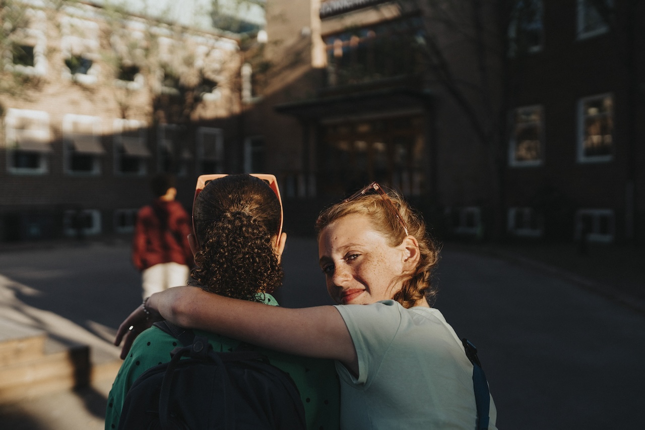 Portrait of smiling girl with arm around friend looking over shoulder while walking at school campus