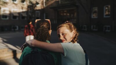 Portrait of smiling girl with arm around friend looking over shoulder while walking at school campus